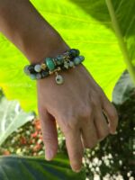 Set di ispirazione con bracciali in pietre naturali: Sfumature di Verde (Turchese africano, Giada verde Birmania, Crisoprasio, Prehinite, Fluorite verde) e bracciale Amazzonite e Agata muschiata indossato su un polso di donna con sfondo la natura illuminata