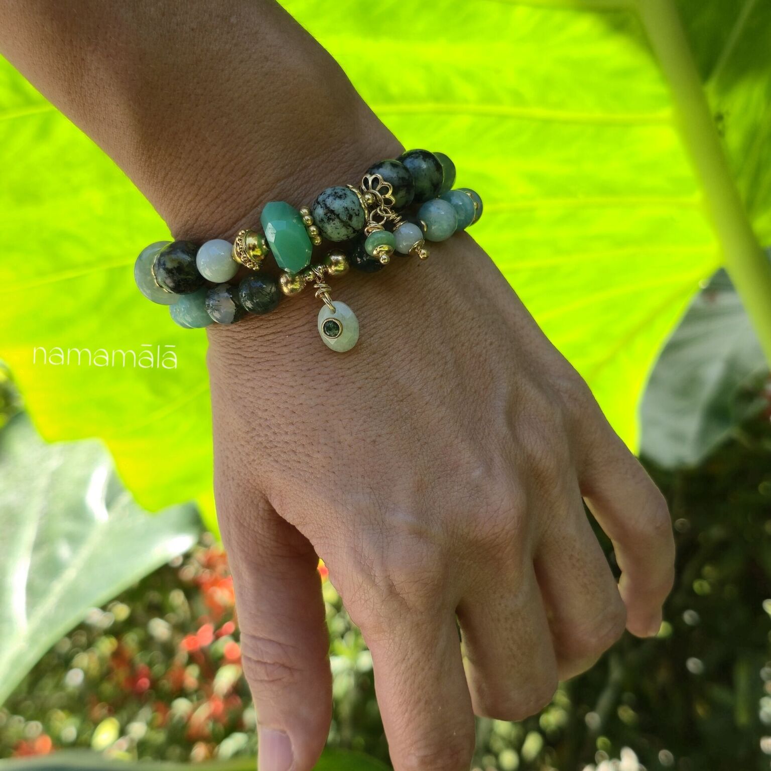 Set di ispirazione con bracciali in pietre naturali: Sfumature di Verde (Turchese africano, Giada verde Birmania, Crisoprasio, Prehinite, Fluorite verde) e bracciale Amazzonite e Agata muschiata indossato su un polso di donna con sfondo la natura illuminata