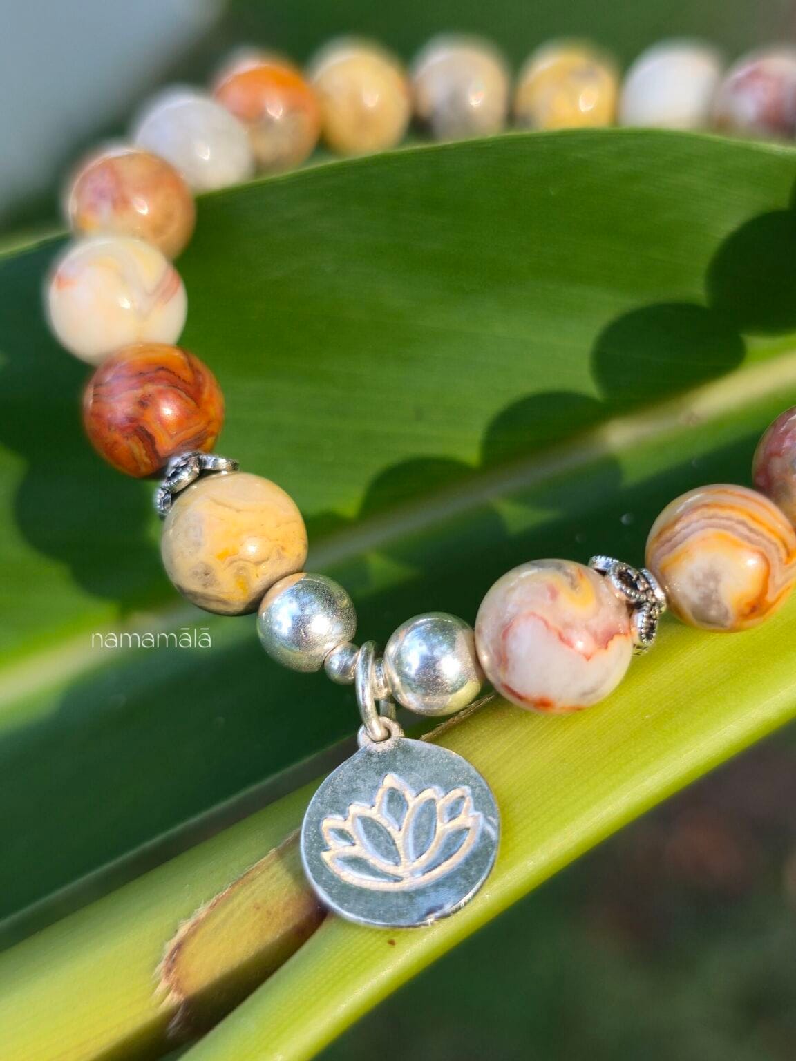 Dettaglio ravvicinato di un bracciale in pietre naturali di agata crazy lace con charm fiore di loto in argento 925 appoggiato su una foglia verde illuminata dal sole.