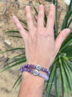Due bracciali in pietre naturali di Ametista mostrati nelle due versioni disponibili: o tutto di colore ametista viola, o in due colori, ametista viola e ametista lavanda, dal colore lilla. Sono indossati su un polso di donna con lo sfondo la natura verde.