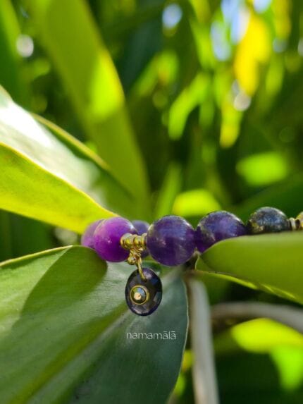 bracciale elastico di ametista e pirite con distanziatori e ciondolo in oro della collezione namamala, posato tra foglie di piante tropicali.