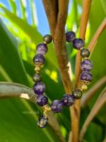 bracciale elastico di ametista e pirite con distanziatori e ciondolo in oro della linea namamala, posato tra foglie di piante tropicali.