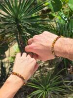 Bracciali di coppia in Occhio di Tigre naturale con distanziatori in argento 925, indossati da un uomo e una donna su sfondo tropicale.