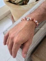 Bracciale in pietre naturali di quarzo ematoide con venature rosse e arancio e charm in argento 925, indossato al polso da un uomo su uno sfondo di legno e piante
