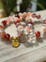 Due bracciali in pietre naturali di quarzo ematoide con venature rosse e arancio, mostrati nelle due versioni con charm e distanziatori in argento 925 e in argento con la placcatura oro, appoggiati su legno chiaro.