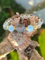 bracciale di pietre naturali appoggiato su legno grezzo con grande zaffiro grezzo grigio-blu al centro affiancato da due perle tonde azzurre di larimar, perle di quarzo trasparente e dettagli placcati oro.