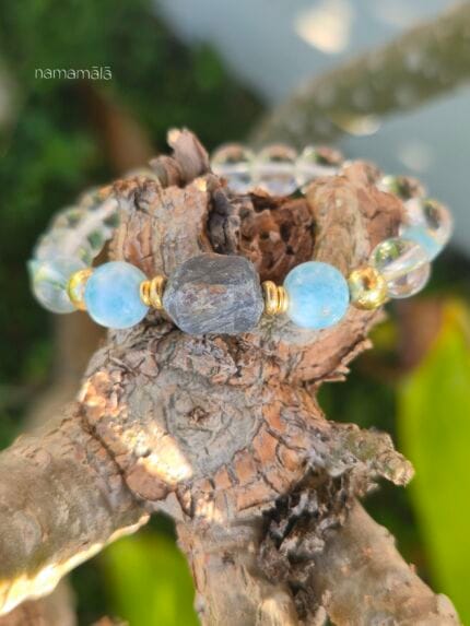 bracciale di pietre naturali appoggiato su legno grezzo con grande zaffiro grezzo grigio-blu al centro affiancato da due perle tonde azzurre di larimar, perle di quarzo trasparente e dettagli placcati oro.