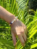 bracciale elastico shoreline con lapislazzuli giada verde calcedonio blu e calcite gialla indossato al polso su sfondo di foglie tropicali