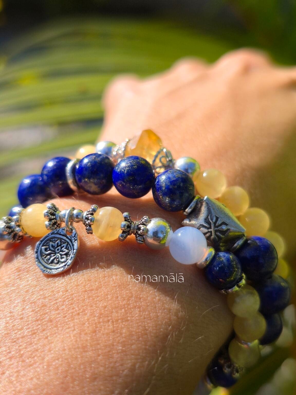 Set di ispirazione con bracciali in pietre naturali: Shoreline (Calcite gialla, Lapislazzuli, Calcedonio blu), Nel Blu dipinto di blu (Lapislazzuli), Manipura (Calcite gialla e Citrino) indossati al polso di donna su sfondo una foglia di palma verdeggiante.