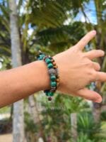 Bracciali da donna con pietre naturali di Occhio di Tigre marrone e Malachite verde con distanziatori in argento 925, indossati al polso e con uno sfondo di palme verdi.