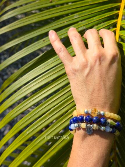 Set di ispirazione con bracciali in pietre naturali: Shoreline (Calcite gialla, Lapislazzuli, Calcedonio blu), Nel Blu dipinto di blu (Lapislazzuli), Manipura (Calcite gialla e Citrino) indossati al polso di donna su sfondo una foglia di palma verdeggiante.