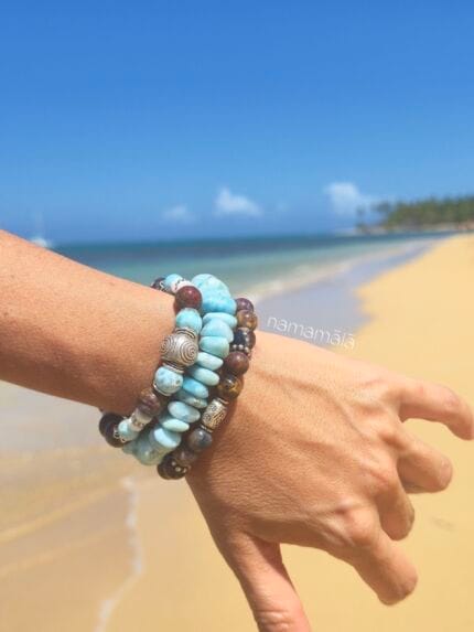 Set di ispirazione con bracciali in pietre naturali: Nuova Energia (tutto in Pietersite), Larimar, Spirale nella Tempesta (Pietersite e Larimar), indossato al mare su una spiaggia su un polso di donna
